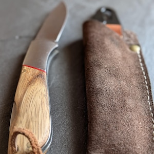 Könnte beinhalten: Ein kleines Messer aus Edelstahl mit Holzgriff und Lederscheide. Das Messer hat eine gebogene Klinge und einen roten Akzent am Griff. Die braune Scheide hat weiße Nähte.