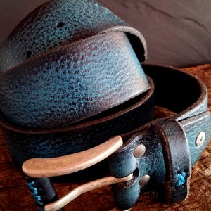 Puede incluir: Un cinturón de cuero envejecido enrollado, con un acabado en color turquesa y marrón. El cinturón presenta una hebilla de color plateado y detalles de costura turquesa. El cuero tiene un aspecto texturizado y el cinturón se muestra sobre una superficie de madera.