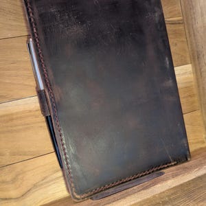 Puede incluir: Una funda rectangular para cuaderno de cuero marrón oscuro con bordes cosidos a mano visibles con hilo oscuro. Un bolígrafo de metal plateado está insertado en una trabilla de cuero unida al lado izquierdo de la funda, junto al lomo.