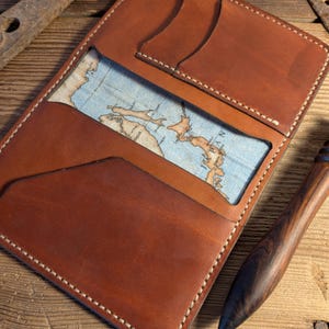 May include: Open tan leather wallet with white stitching and a map-printed fabric interior. The wallet has multiple card slots and a rectangular shape. A wooden pen is next to the wallet.