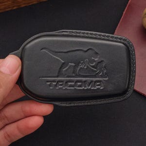 May include: Black leather key fob with a debossed dinosaur design and the word "TACOMA" below. The fob is rectangular with rounded edges and a small loop for attaching to a keychain. The leather has a matte finish.