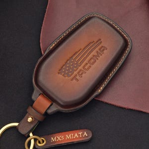 May include: A brown leather key fob with a gold key ring and a small tag that says "MX5 MIATA." The key fob has the word "TACOMA" and a graphic of the American flag. The key fob is made of brown leather and has a stitched border.