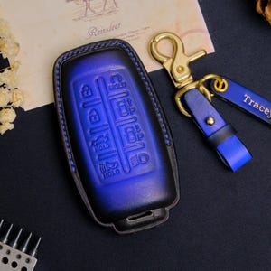 May include: A blue and black leather car key fob cover with embossed buttons. A matching blue leather keychain with a gold-tone clasp and a black tag with the name "Tracey" is attached. The key fob cover is next to a vintage postcard.
