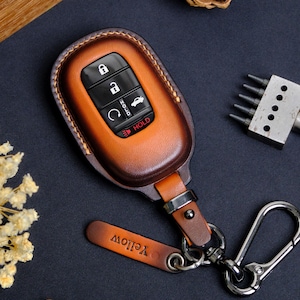 May include: A brown leather car key fob cover with a key ring and a small leather tag that says "Yellow". The key fob has black buttons for locking, unlocking, and other functions. The key fob is a warm brown color.