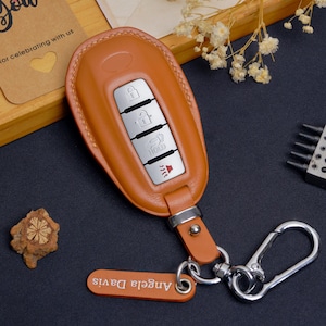 May include: A tan leather car key fob cover with silver buttons and a keychain. The fob has a leather strap and a small tag with the name "Angela Davis". The key fob is on a dark surface.