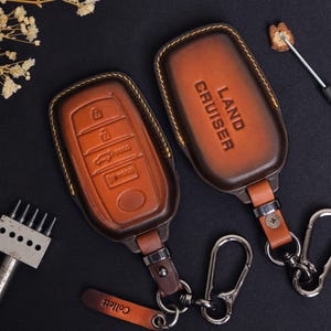 May include: Two brown leather key fob covers with yellow stitching. One cover has buttons for key functions, the other says "LAND CRUISER". Both have key rings and a small leather tag that says "Collett".