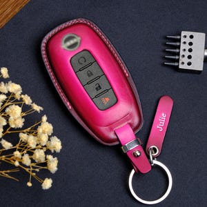 Może przedstawiać: Magenta skórzane etui na kluczyk samochodowy z pasującym brelokiem. Brelok ma trzy przyciski i logo samochodu. Na breloku wygrawerowano imię "Julie".