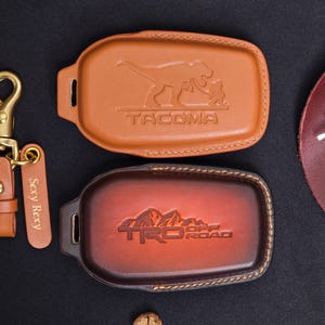 May include: Two brown leather key fob covers with embossed designs. One features a dinosaur and the word "TACOMA", the other has "TRD OFF ROAD" with a mountain graphic. A matching leather keychain with "Sexy Rexy" is also included.