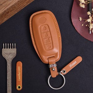 May include: A tan leather car key fob cover with a key ring and a small tag that says "Nissan". The cover has embossed key symbols. A metal tool and a small leather tag with "Leather1993" are also in the image.