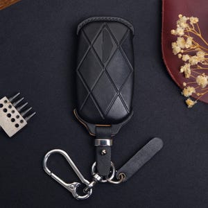 May include: Black leather key fob cover with a diamond pattern and silver-tone hardware. The key fob has a strap and a metal carabiner clip. The background is dark with a leather piece and a sprig of dried flowers.