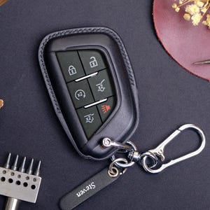 May include: Black leather car key fob cover with silver-tone hardware. The key fob has buttons for locking, unlocking, and other vehicle functions. A small leather tag with the name "Steven" is attached to the key ring.