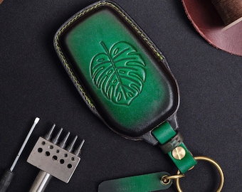 May include: Green leather key fob with a monstera leaf design. The key fob has a brass ring and a small green tag. The edges are stitched with yellow thread. The key fob is on a black surface.