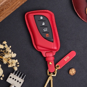 May include: Red leather car key fob cover with a gold-colored clasp and a matching tag with the initials "BHK". The key fob has black buttons and a "HOLD" button. The cover is designed to protect the car key.