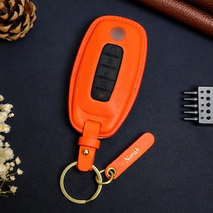 May include: An orange leather key fob cover with a matching strap and gold-colored keyring. The fob has black buttons and a small, engraved tag that reads "Norah".
