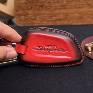 May include: Red leather key fob cover with the Toyota Supra logo embossed on the front. The cover has a dark brown border and red stitching. A silver-colored metal ring and leather strap are attached.