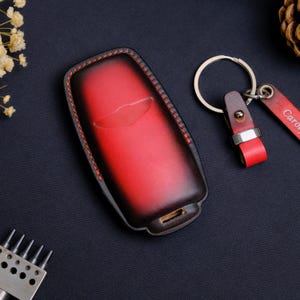May include: A red and black gradient leather key fob cover with red stitching and a matching leather keychain strap featuring a silver metal ring. The key fob cover has a molded shape with a central indentation and a small rectangular opening at the base.