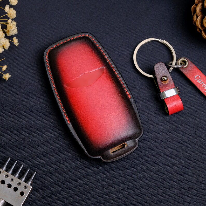 May include: A red and black gradient leather key fob cover with red stitching and a matching leather keychain strap featuring a silver metal ring. The key fob cover has a molded shape with a central indentation and a small rectangular opening at the base.