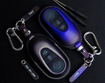 May include: Two car key fobs in dark purple and blue leather cases with keychains. One fob has a tag that says "Starr". A small tag with the phone number 828-301-2298 is also present. Two small screwdrivers are in the background.