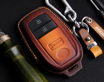 May include: A brown leather key fob with a silver key ring and a black car key inside. The key fob has a stitched edge and a small button on the bottom. The key ring has a small metal tag with the word "BREH" on it.