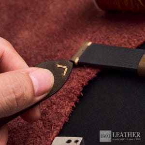 May include: Close-up of a leather craft project. A small, dark brown leather piece with a stitched corner is held near a black leather strap. The image shows the process of leather crafting, with a spool of thread in the background.