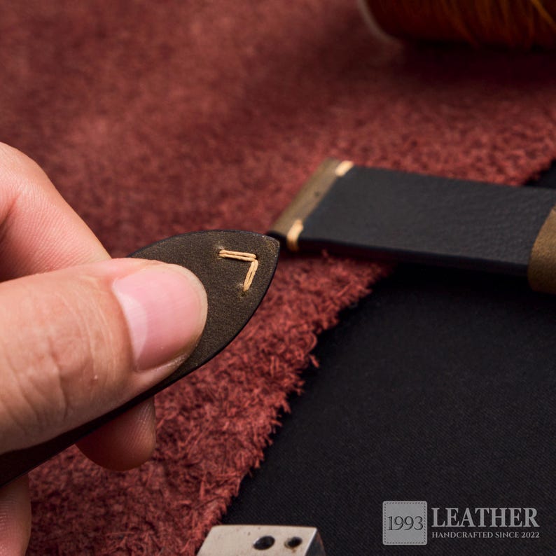 May include: Close-up of a leather craft project. A small, dark brown leather piece with a stitched corner is held near a black leather strap. The image shows the process of leather crafting, with a spool of thread in the background.