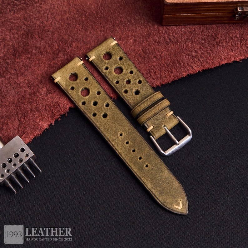 Puede incluir: Correas de reloj de cuero verde oliva con m&uacute;ltiples perforaciones circulares. Las correas tienen un aspecto vintage, con costuras visibles y una hebilla plateada. El texto "1993 LEATHER HANDCRAFTED SINCE 2022" es visible en la parte inferior.
