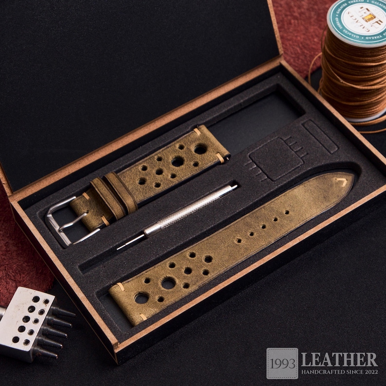 Puede incluir: Un juego de correas de reloj en una caja de madera. El juego incluye una correa de reloj de cuero verde oliva desgastado con agujeros perforados, una herramienta plateada y una segunda pieza de correa. La caja est&aacute; forrada con espuma negra. El texto "1993 LEATHER HANDCRAFTED SINCE 2022" es visible.