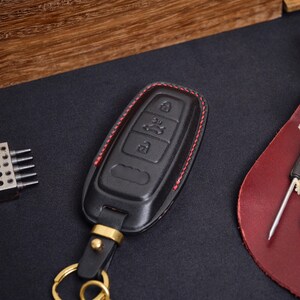 May include: A black leather car key fob cover featuring red stitching along the edges. The cover has embossed icons for locking, unlocking, and trunk release functions. It includes a gold-toned metal attachment loop and a circular key ring.