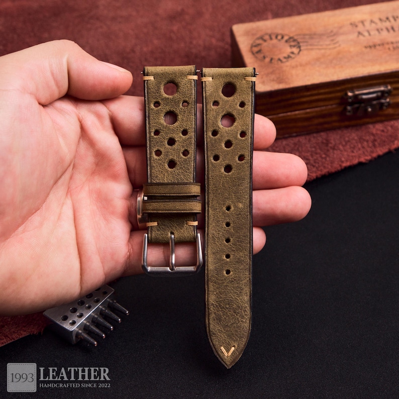 Puede incluir: Correas de reloj de cuero verde oliva con agujeros perforados y costuras en contraste. Las correas tienen una hebilla plateada y est&aacute;n estampadas con "1993 LEATHER HANDCRAFTED SINCE 2022".