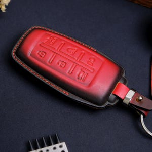 May include: A red and black gradient leather key fob cover featuring embossed icons for lock, unlock, trunk release, and remote start functions. The cover is stitched with red thread and includes a matching leather strap with a silver metal ring.