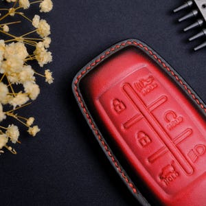 May include: A red and black leather car key fob cover featuring embossed button icons for locking, unlocking, trunk release, and panic alarm functions. The cover is stitched with red thread along the edges and has a smooth, gradient finish.