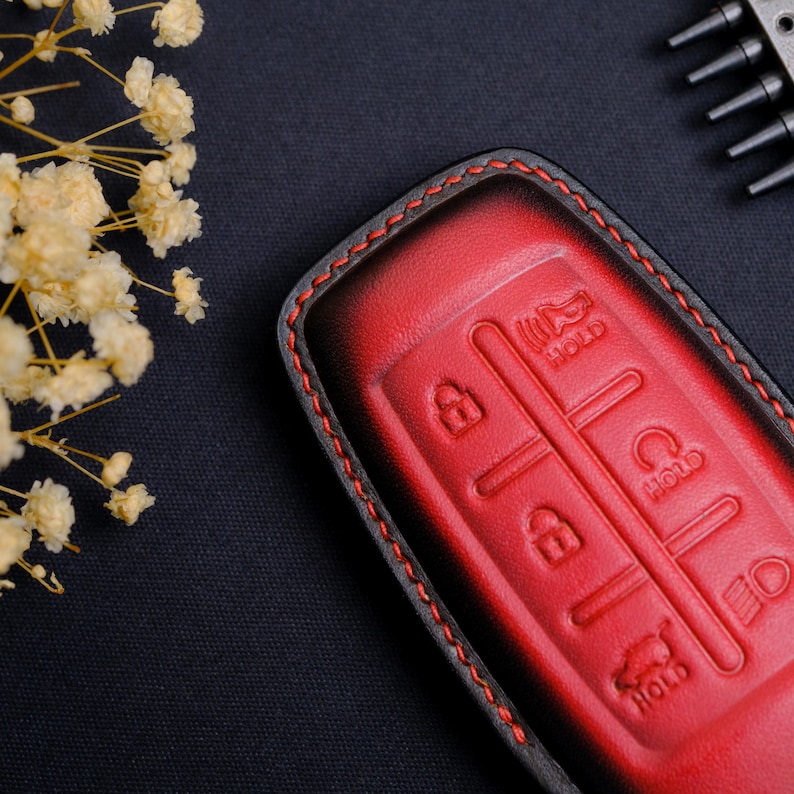 May include: A red and black leather car key fob cover featuring embossed button icons for locking, unlocking, trunk release, and panic alarm functions. The cover is stitched with red thread along the edges and has a smooth, gradient finish.