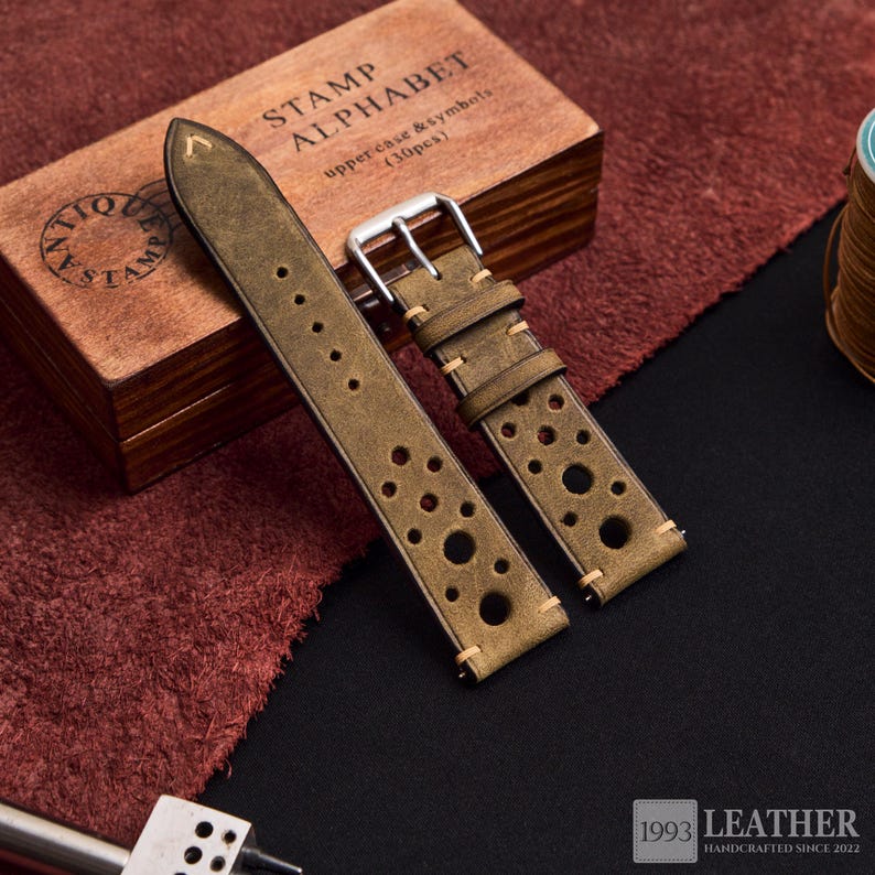 Puede incluir: Correa de reloj de cuero verde oliva con agujeros perforados y costuras color crema. La correa tiene una hebilla plateada y se muestra con una caja de madera de sellos de alfabeto. El texto "1993 LEATHER HANDCRAFTED SINCE 2022" es visible.
