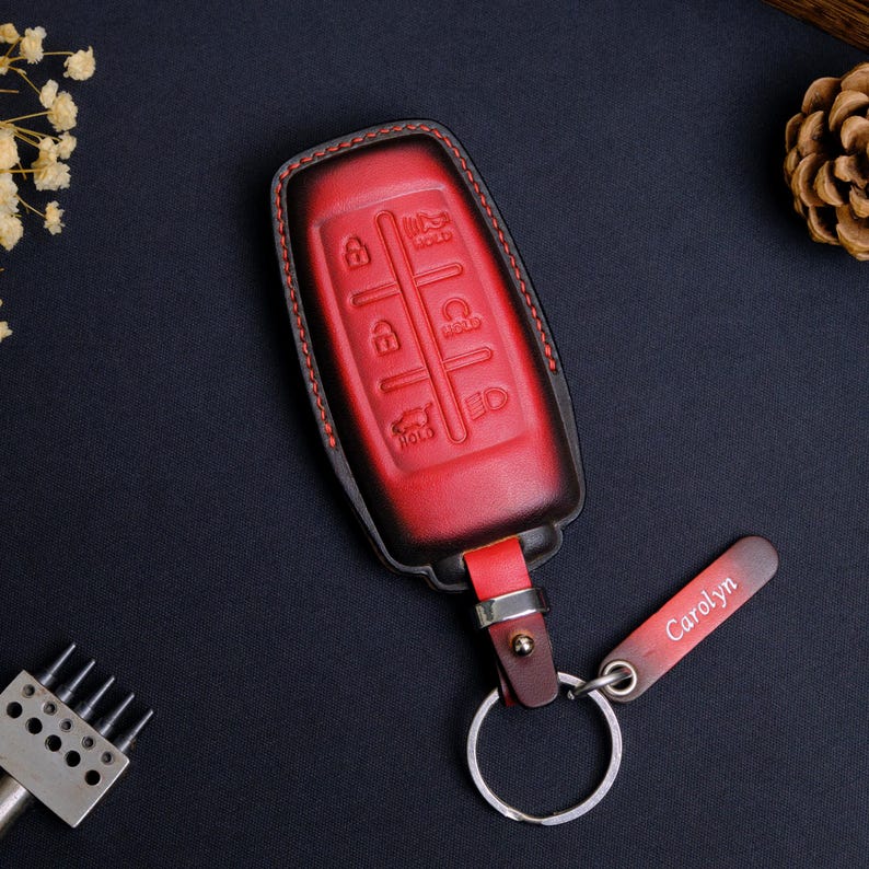 May include: A red and black gradient leather car key fob cover featuring embossed button icons for lock, unlock, trunk, and remote start functions. The cover is attached to a silver metal keyring with a small rectangular leather tag engraved with the name Carolyn.