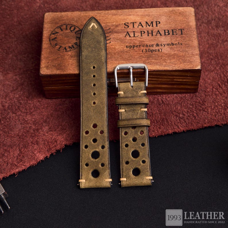 Puede incluir: Correas de reloj de cuero verde oliva con perforaciones circulares y costuras crema. Se muestran junto a una caja de madera con el texto "STAMP ALPHABET" y "upper case & symbols (30pcs)". La imagen tambi&eacute;n incluye el texto "1993 LEATHER HANDCRAFTED SINCE 2022".