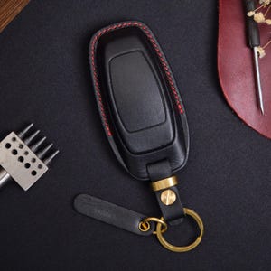 May include: A black leather car key fob cover featuring red contrast stitching along the edges. The cover is attached to a brass-coloured metal ring and a small black leather tag, secured by a flat-head screw and a brass-coloured metal loop.