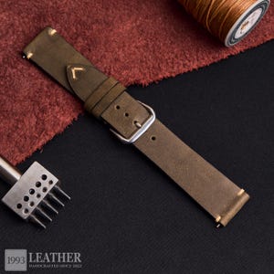 May include: Olive green leather watch strap with a silver buckle. The strap is stitched with cream-colored thread. The image also shows a spool of thread, a leather punch tool, and a piece of red fabric. The text "1993 LEATHER HANDCRAFTED SINCE 2022" is visible.