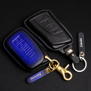 May include: Two leather key fob covers, one black and one blue, with keychains. The black cover has embossed key symbols. The blue cover has embossed key symbols and the name "Jeannine" on the keychain. The black keychain says "Lincoln".