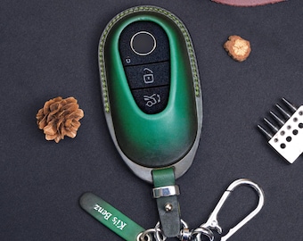May include: An emerald green leather key fob cover with a black plastic remote insert featuring three buttons. The cover has green stitching and is attached to a silver metal carabiner and a small green leather tag with the text Ki's Benz printed.