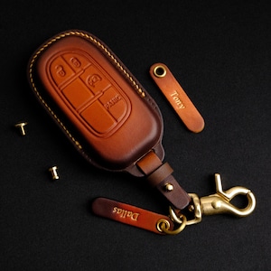 May include: A brown leather key fob with a metal key ring and a separate leather tag that says "Dallas". The key fob has a stitched edge and a metal button that says "Panic".