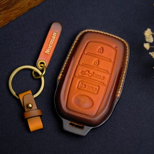 May include: A brown leather car key fob cover and matching keychain. The key fob cover has embossed buttons and a key symbol. The keychain has a gold ring and the name "Norman" embossed on the leather tag.