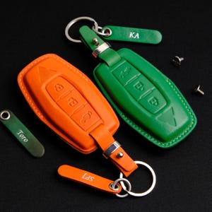 May include: Two leather key covers for car keys. One is orange and the other is green. Both have a key ring and a small leather tag with the text "KA" and "SPT" on them.