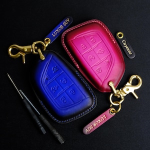 May include: Two leather key fob covers, one blue and one pink, with gold-colored hardware. The blue cover has the text "LUXOR SUV" on a matching blue tag. The pink cover has a matching tag with the text "Cayenne".