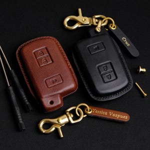 May include: Two leather key covers, one brown and one black, with gold metal keychains. The brown keychain has a tag that says "Yessica Vazpuex" and the black keychain has a tag that says "Cruz".