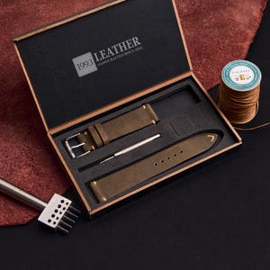 May include: A watch strap kit in a wooden box. The kit includes a dark olive green leather watch strap, a silver tool, and a buckle. The box is lined with black material and has the text "1993 LEATHER HANDCRAFTED SINCE 2022". A spool of thread is also visible.