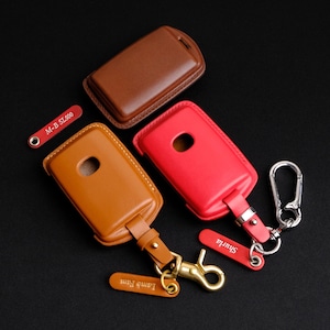May include: Three leather keychains with different colored key covers. The key covers are brown, tan, and pink. Each keychain has a metal clasp and a small tag with text. The tags read "M-B SL500", "Lamb Farm", and "Shrutila".