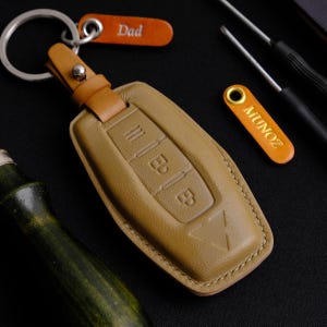 Może przedstawiać: Brązowy skórzany brelok do kluczy z beżowym paskiem ze skóry i srebrnym kółkiem na klucze. Brelok ma trzy przyciski, a na małej beżowej skórzanej metce przymocowanej do kółka na klucze wygrawerowano napis "Dad".