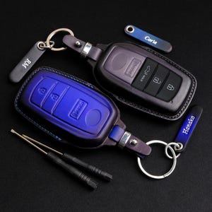 Puede incluir: Dos llaveros de cuero con nombres personalizados, "Carlie" y "Hanako", unidos a fundas para llaves de coche. Una funda para llaves es de cuero negro con una llave plateada, y la otra es de cuero azul con una llave plateada.