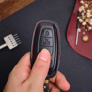 May include: A black leather car key fob cover featuring red contrast stitching along the edges. The cover has moulded button icons for locking, unlocking, and trunk release functions, with a metal attachment loop at the bottom base.