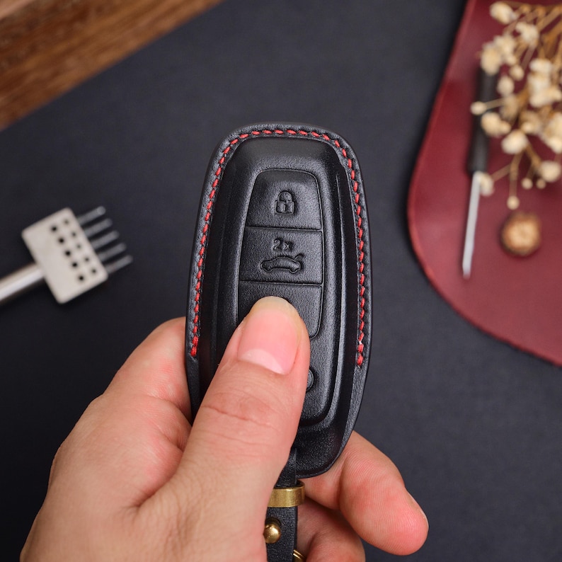 May include: A black leather car key fob cover featuring red contrast stitching along the edges. The cover has moulded button icons for locking, unlocking, and trunk release functions, with a metal attachment loop at the bottom base.